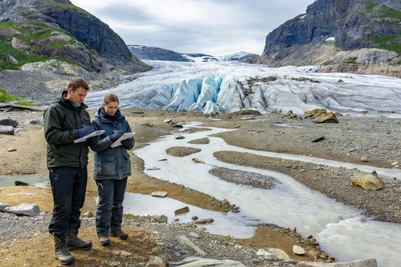 Two field observers with notebooks looking across a braided glacial meltwater stream toward the crevassed snout of a Norwegian valley glacier