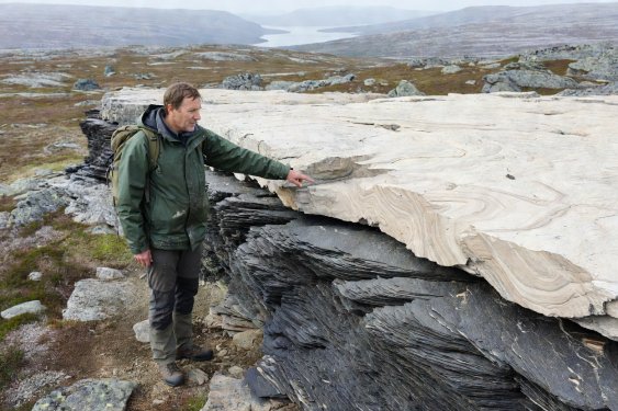 Hiker pointing at a sharp horizontal geological contact between dark flaky phyllite below and pale massive gneiss above in Norway